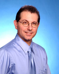 Image of dental professional instructor in blue shirt and blue tie with glasses on looking in the direction of the camera.